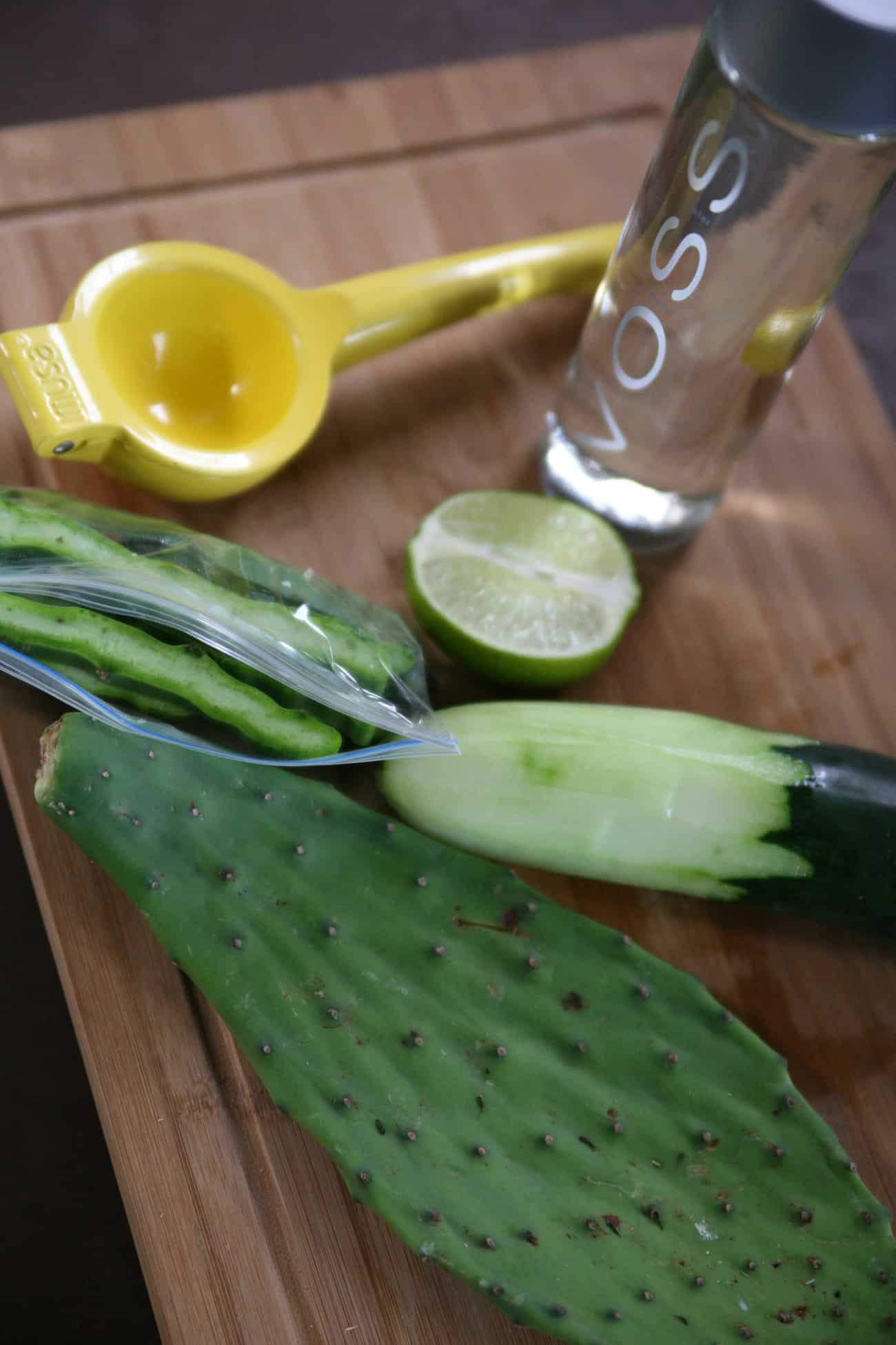 bottle of voss water, a nopal pad, a cucumber that is half peeled, a lemon juicer and a halved lime on a cutting board for making cactus smoothies (licuado de nopal).