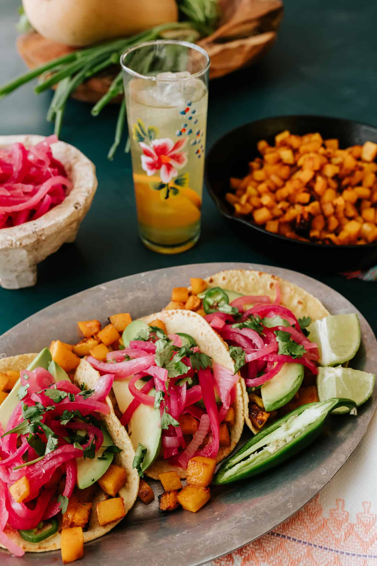 silver serving platter with three roasted butternut squash tacos topped with pickled red onions on a table with a floral painted glass filled with an orange soda water spritz.