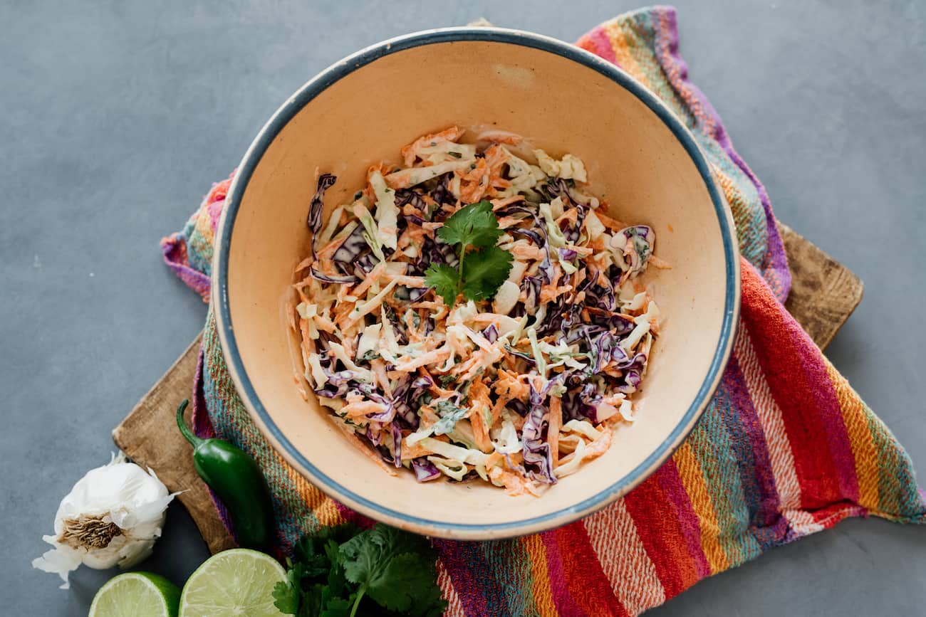 bowl of spicy coleslaw for fish tacos in a bowl on a striped multi-colored tea towel with a head of garlic, halved lime, bunch of cilantro and whole jalapeño on the side.