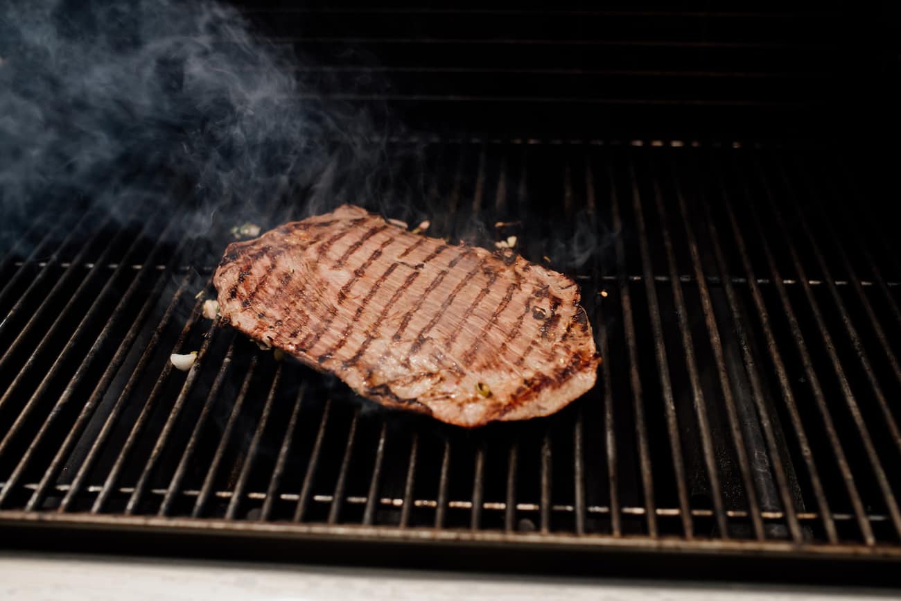 Flank steak (carne asada) on a grill with smoke in the background.