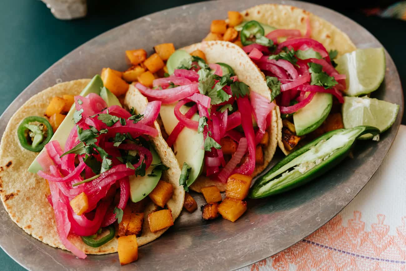 close up of an oblong silver serving platter with an assortment of butternut squash tacos topped with pickled red onions, fresh cilantro, creamy avocado slices and sliced jalapeños.