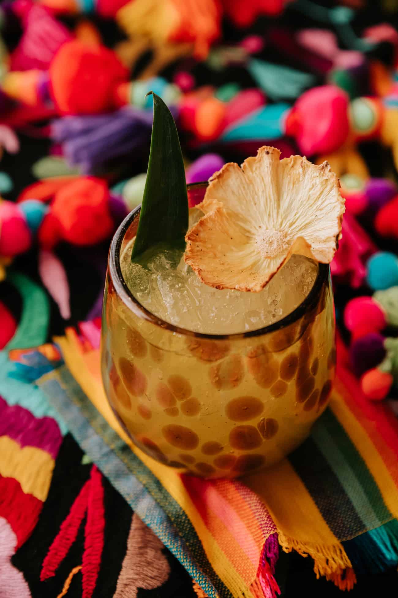 Flor de Pina cocktail in a speckled amber glass on a colorful patterned background garnished with a dried pineapple flower and a pineapple leaf.