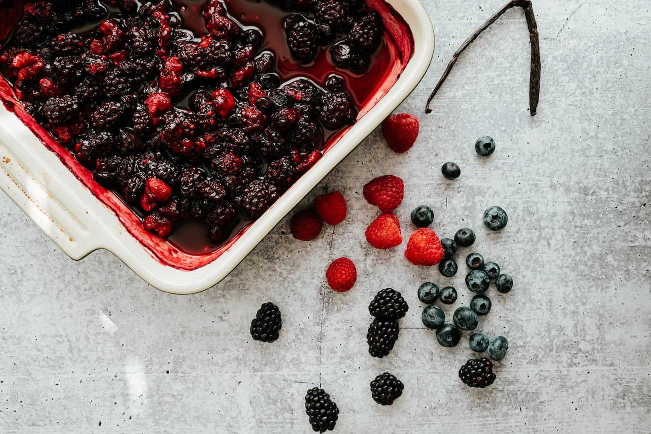 berries in a white roasting pan with some fresh berries and a vanilla bean on the side.