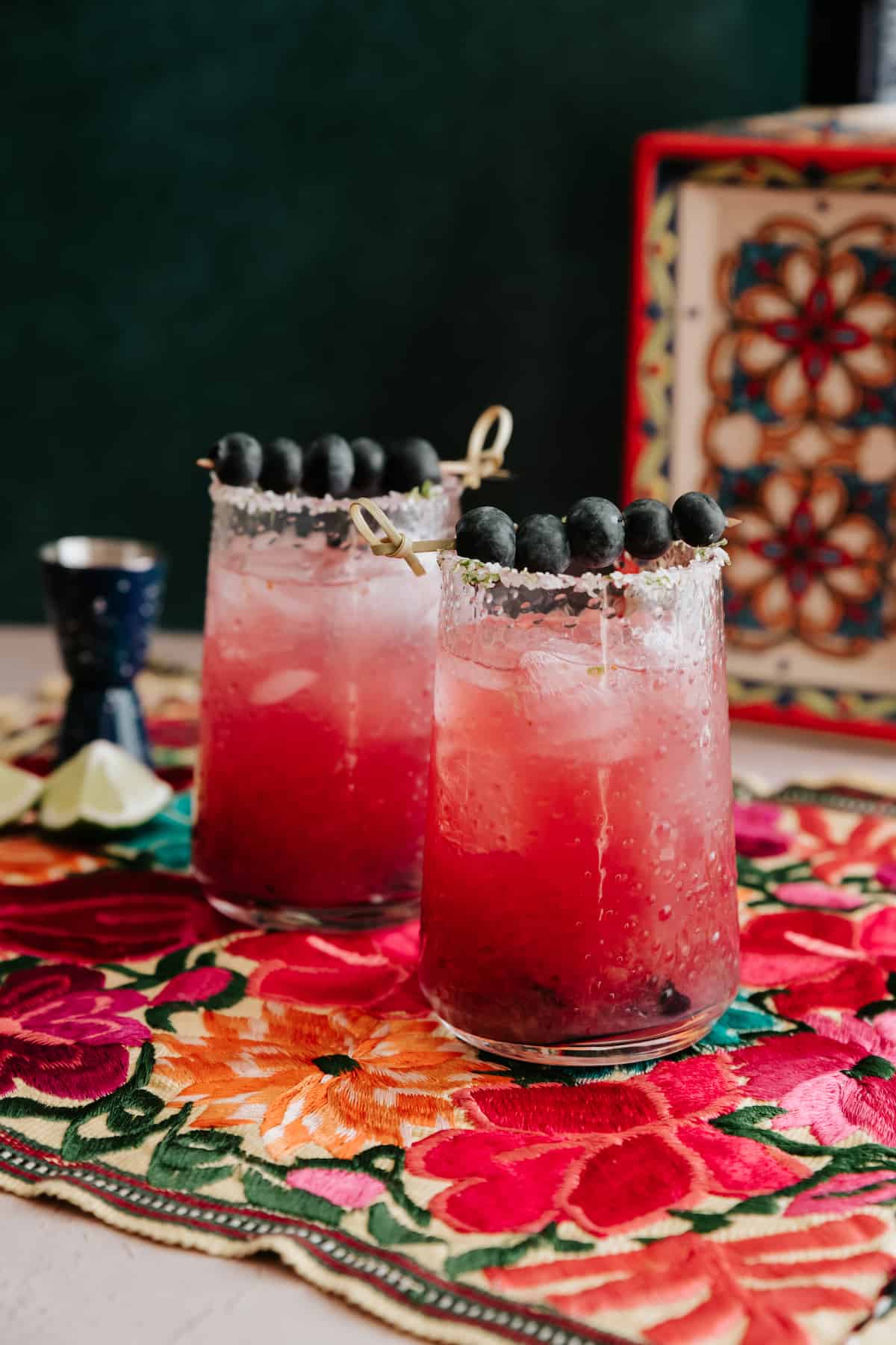 two blueberry margaritas adorned with toothpicks of blueberries on a floral printed tablecloth.