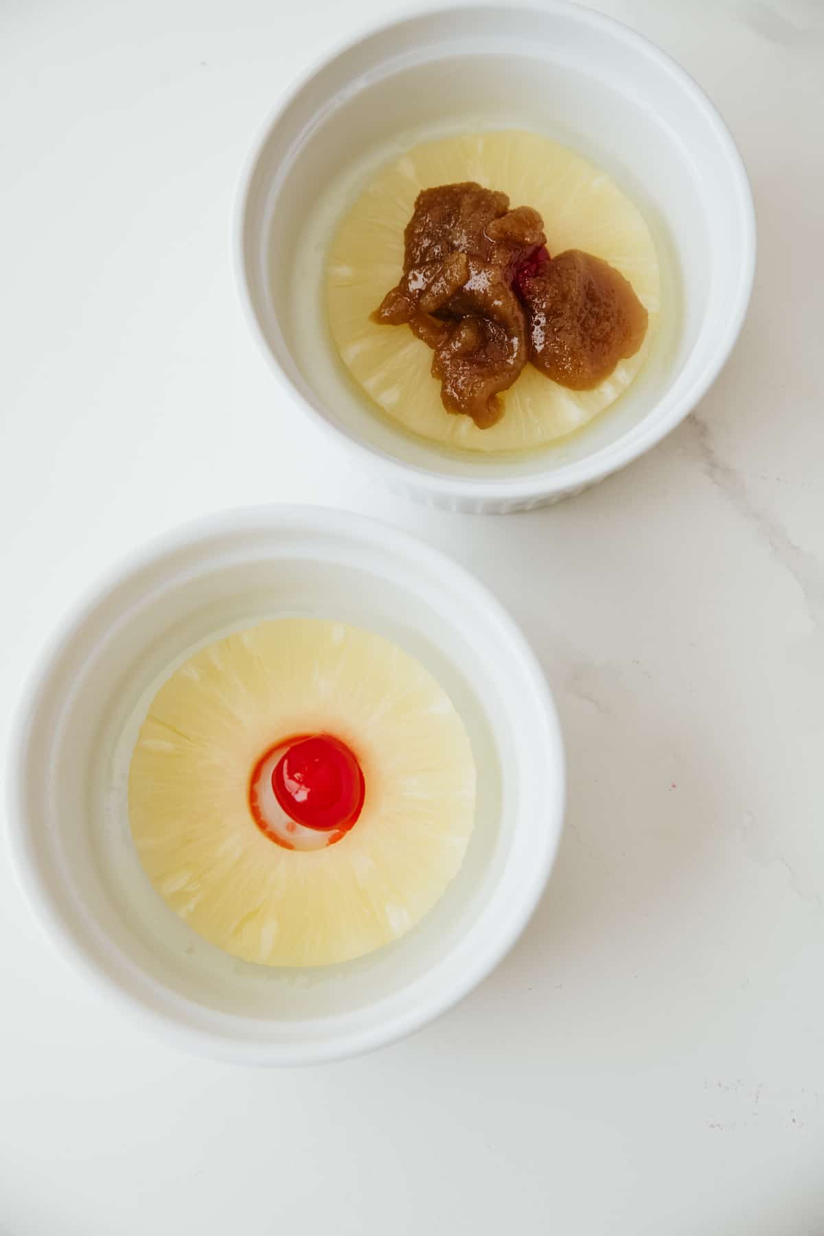 close up of two ramekins with pineapple and maraschino cherries, one of which has the brown sugar caramel topping on top.