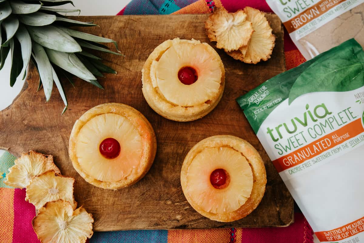 overhead shot of 3 mini pineapple upside down cakes on a wooden cutting board with bags of truvia sweet complete off to the side.