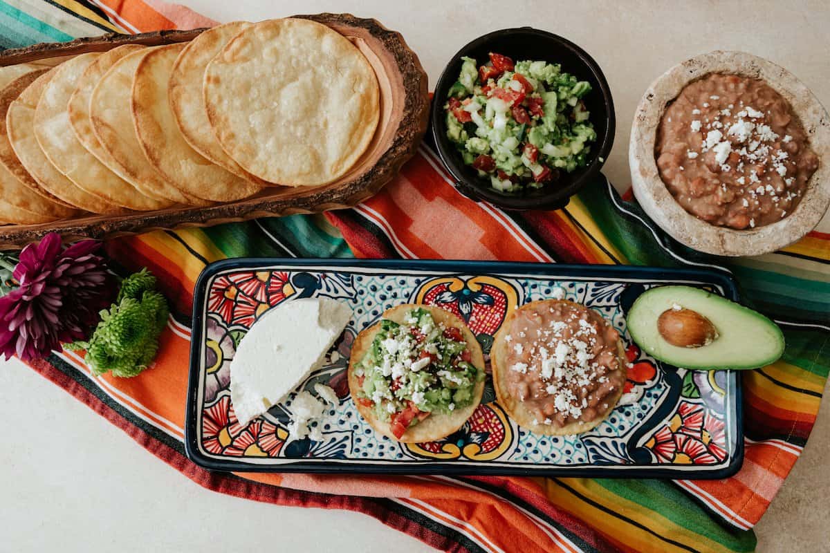 Vegetarian Mexican Tostadas With Refried Beans & Guacamole on a Mexican talavera platter on a striped fabric with bowls on toppings and tostadas on the table.
