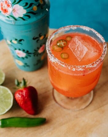 strawberry jalapeno margarita in a clear footed glass with a turquoise floral cocktail shaker in the background.