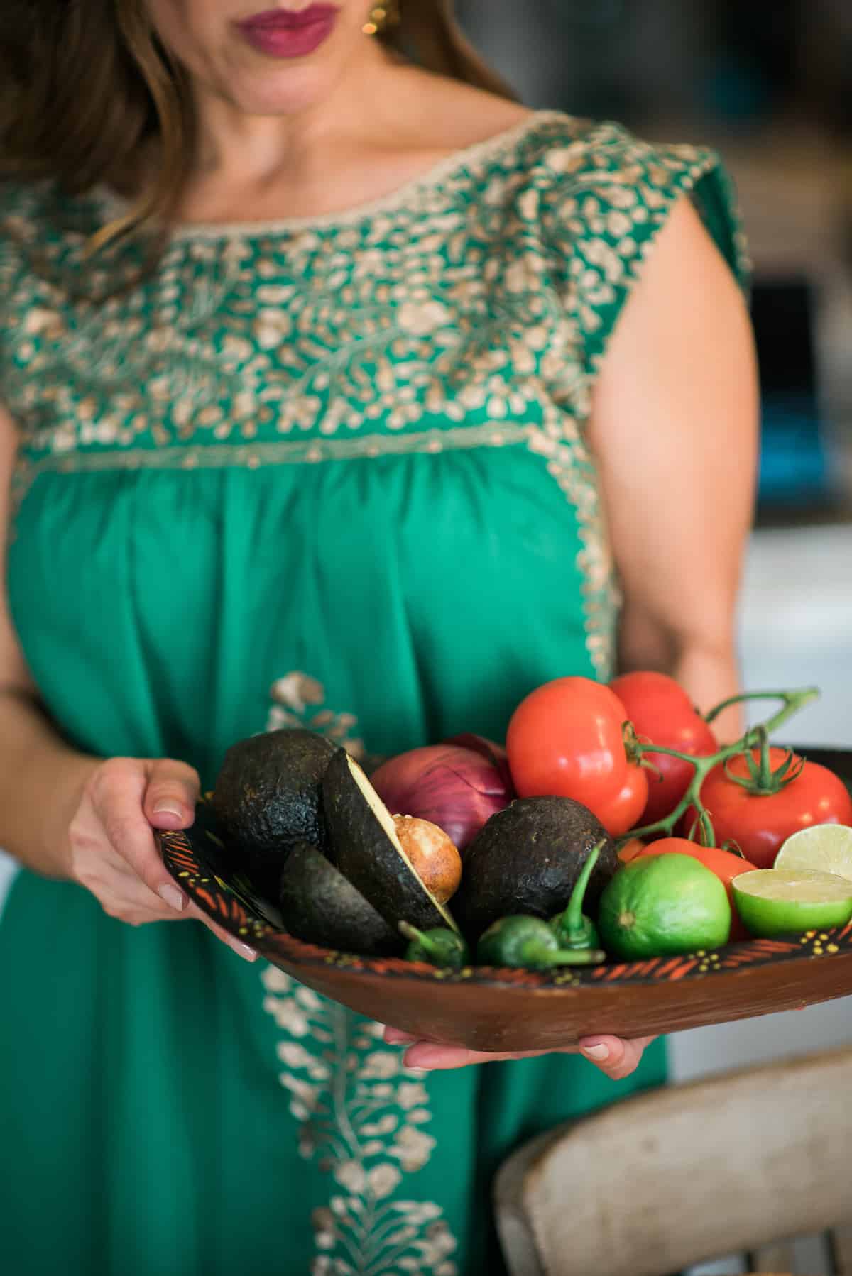 latina blogger holding a wooden bowl filled with tomatoes on the vine, fresh limes, red onion, jalapenos, and avocado for making traditional guacamole.