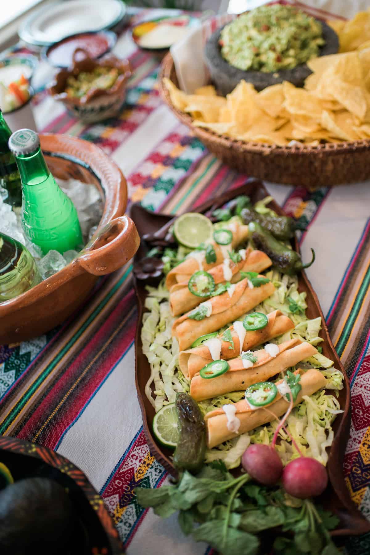 overhead shot of a serving tray of mexican flautas filled with pork tinga on a table with a bucket of beer and a bowl of chips and guacamole.