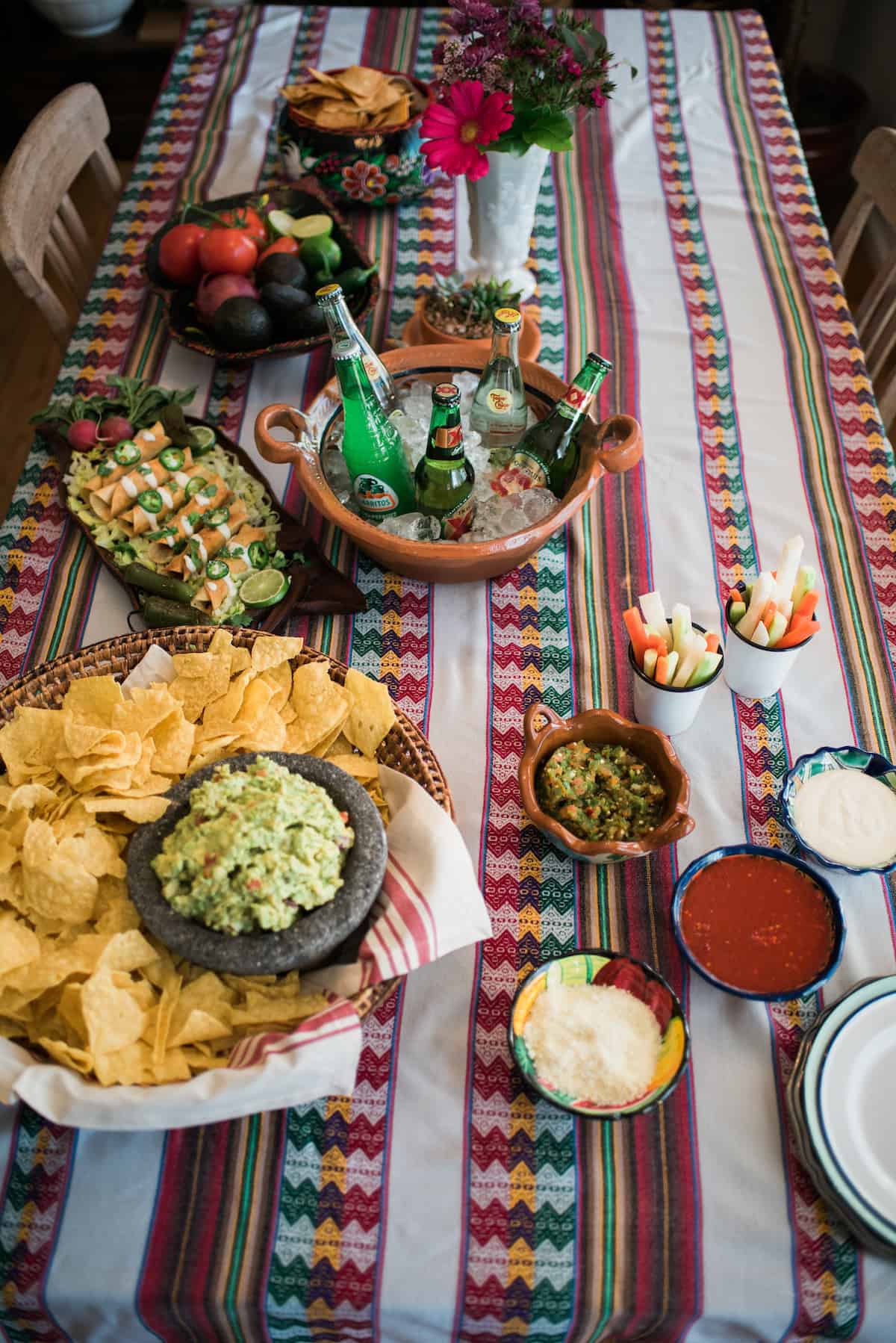 overhead shot of a party spread with chips, guacamole, flautas, veggie cups, salsas, sour cream, cotija cheese, and beer.