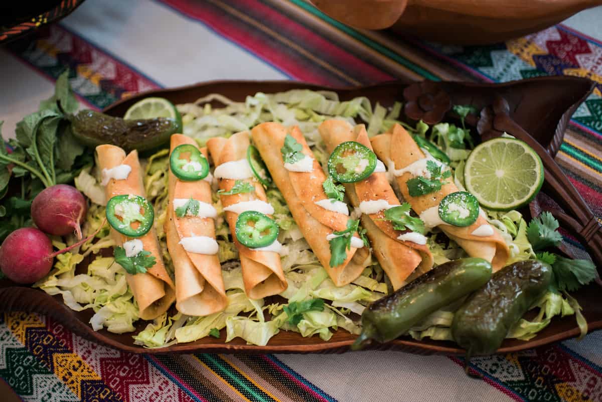 Mexican pork flautas (also known as taquitos or tacos dorados) on a bed of sliced lettuce, drizzled with crema, sprinkled with fresh jalapeño slices and cilantro leaves, with some grilled whole jalapenos on the platter.