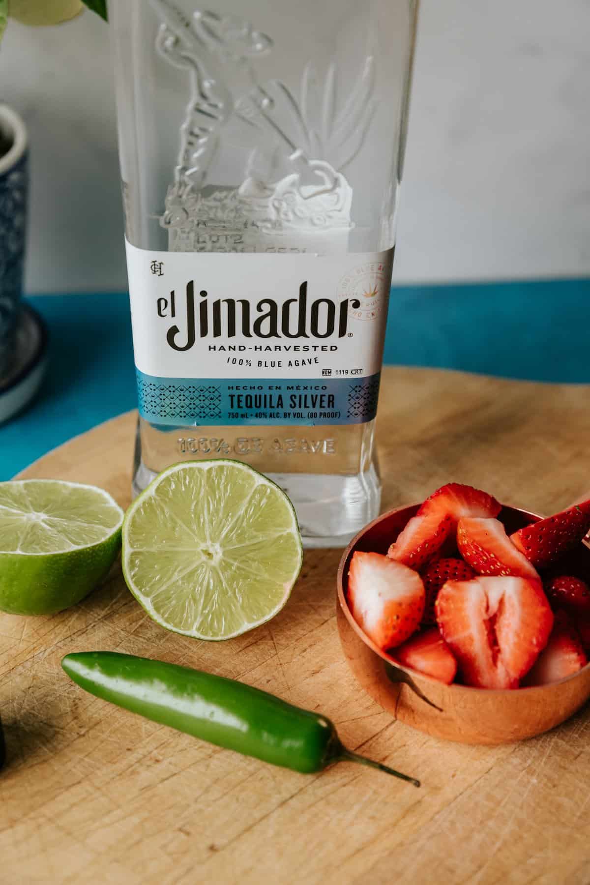 bottle of el jimador tequila blanco on a wooden cutting board with a halved lime, some sliced strawberries, and a serrano pepper.