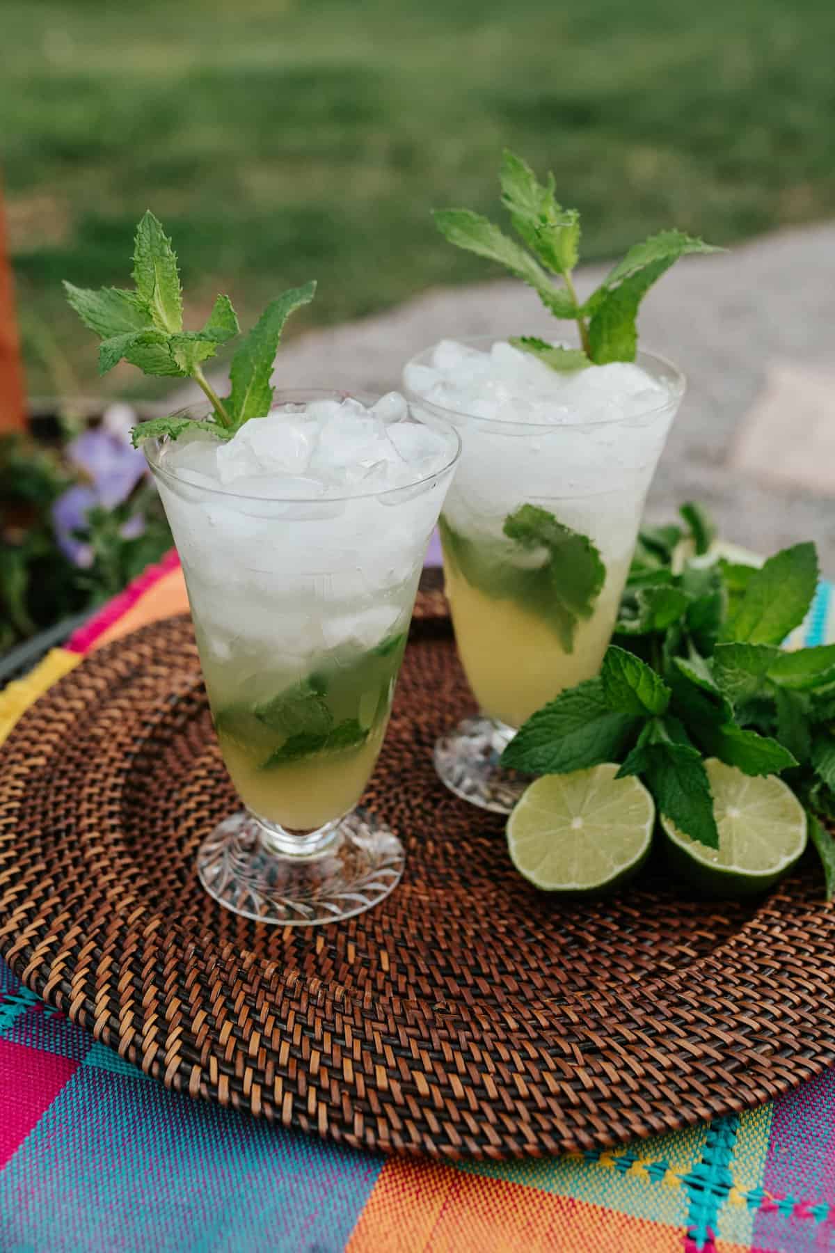 two hurricane glasses filled with classic mint mojitos on a rattan placemat with fresh mint and halved limes on the side. 