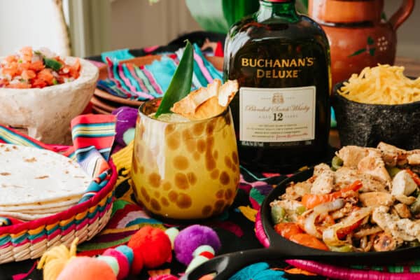 flor de pina cocktail on a table with a bottle of buchanan's whisky and a skillet of chicken fajitas.