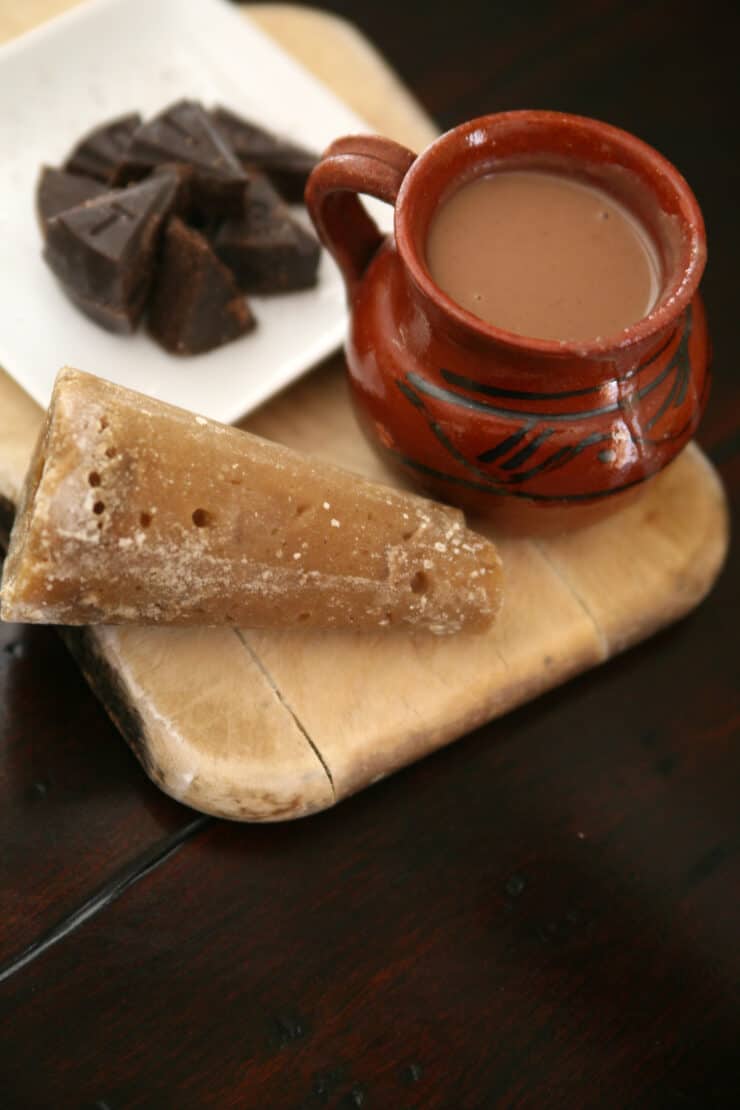 cone of piloncillo on a cutting board with a mug of mexican hot chocolate and a circle of mexican chocolate cut into wedges.