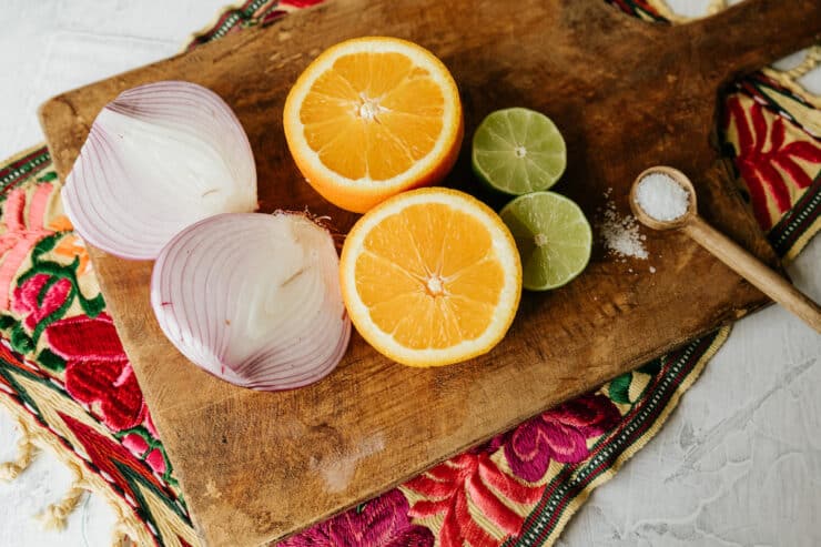 a halved red onion, a halved orange, a halved lime, and a wooden spoon of salt on a wooden cutting board.