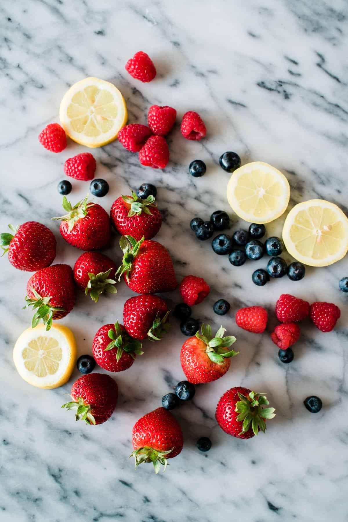 fresh strawberries, raspberries, blueberries, and lemon slices strewn across a marble counter. 