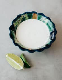 blue and green painted scalloped bowl filled with homemade crema mexicana on a grey table with a few fresh lime wedges to the side.