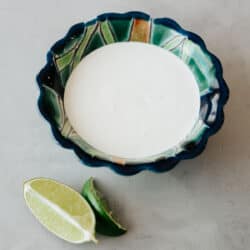 Cuenco festoneado pintado de azul y verde lleno de crema mexicana casera sobre una mesa gris con unos cuantos gajos de lima fresca a un lado.