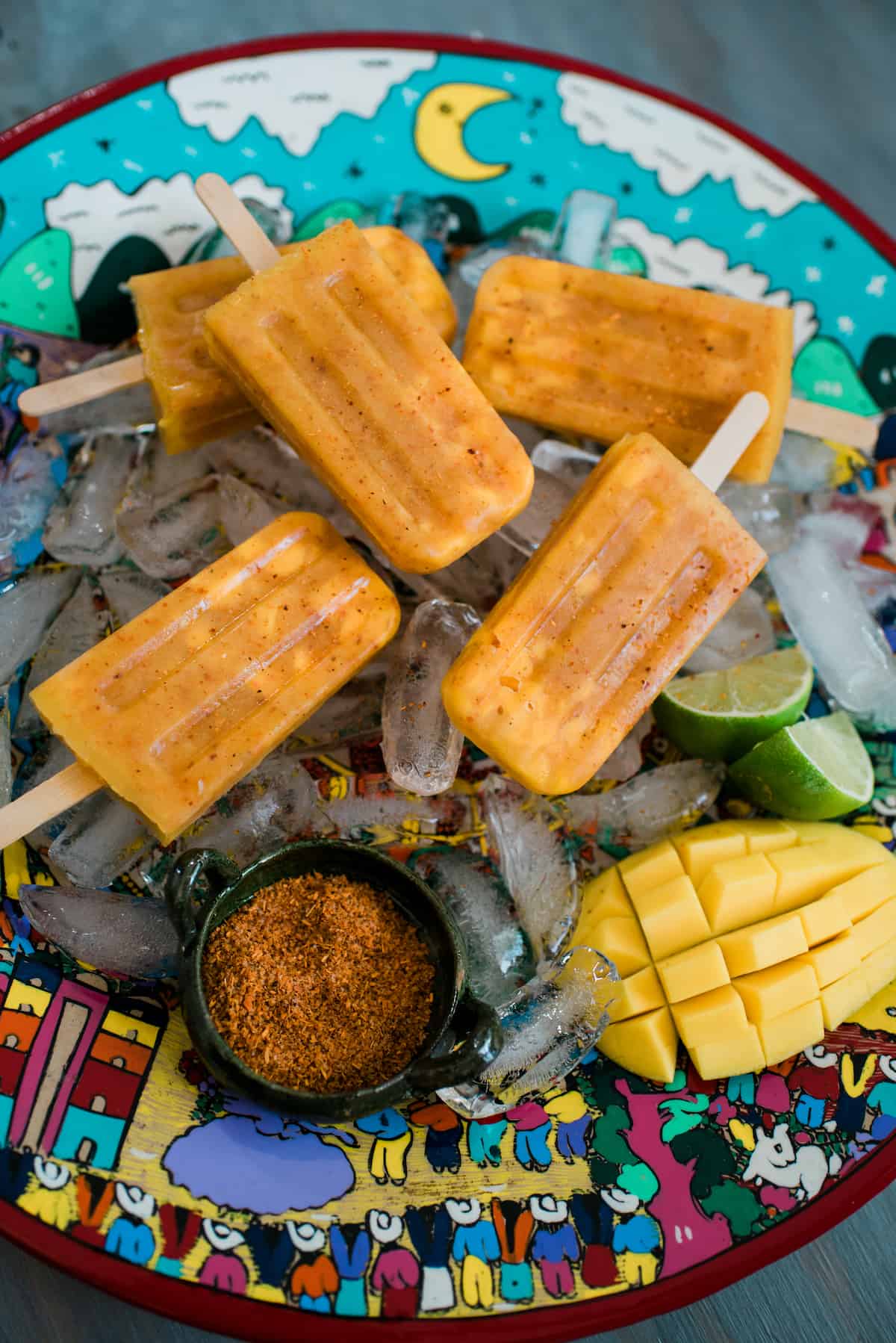 serving platter of chili mango paletas on a bed of ice with a ramekin of tajin and a halved lime.