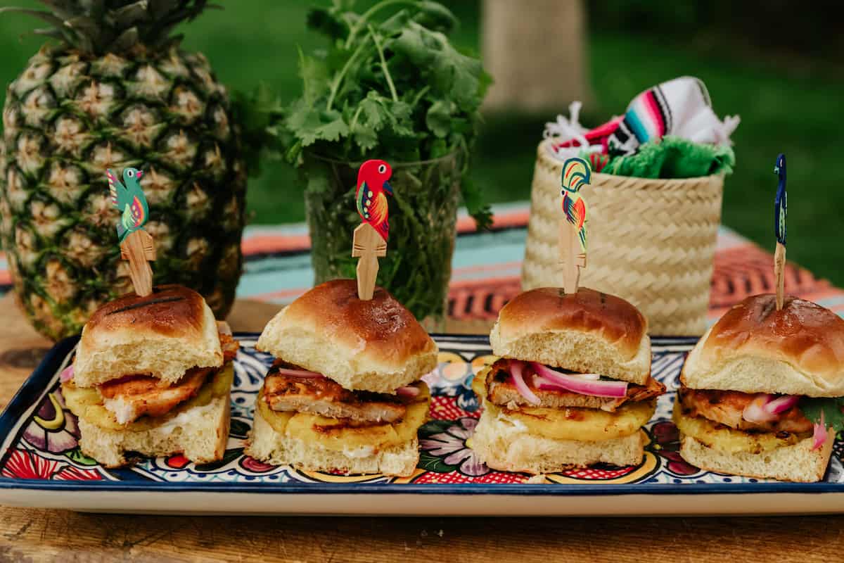 tablescape of Mexican pork chop sliders al pastor with a bouquet of fresh cilantro, a whole pineapple, and a wicker basket of napkins. 