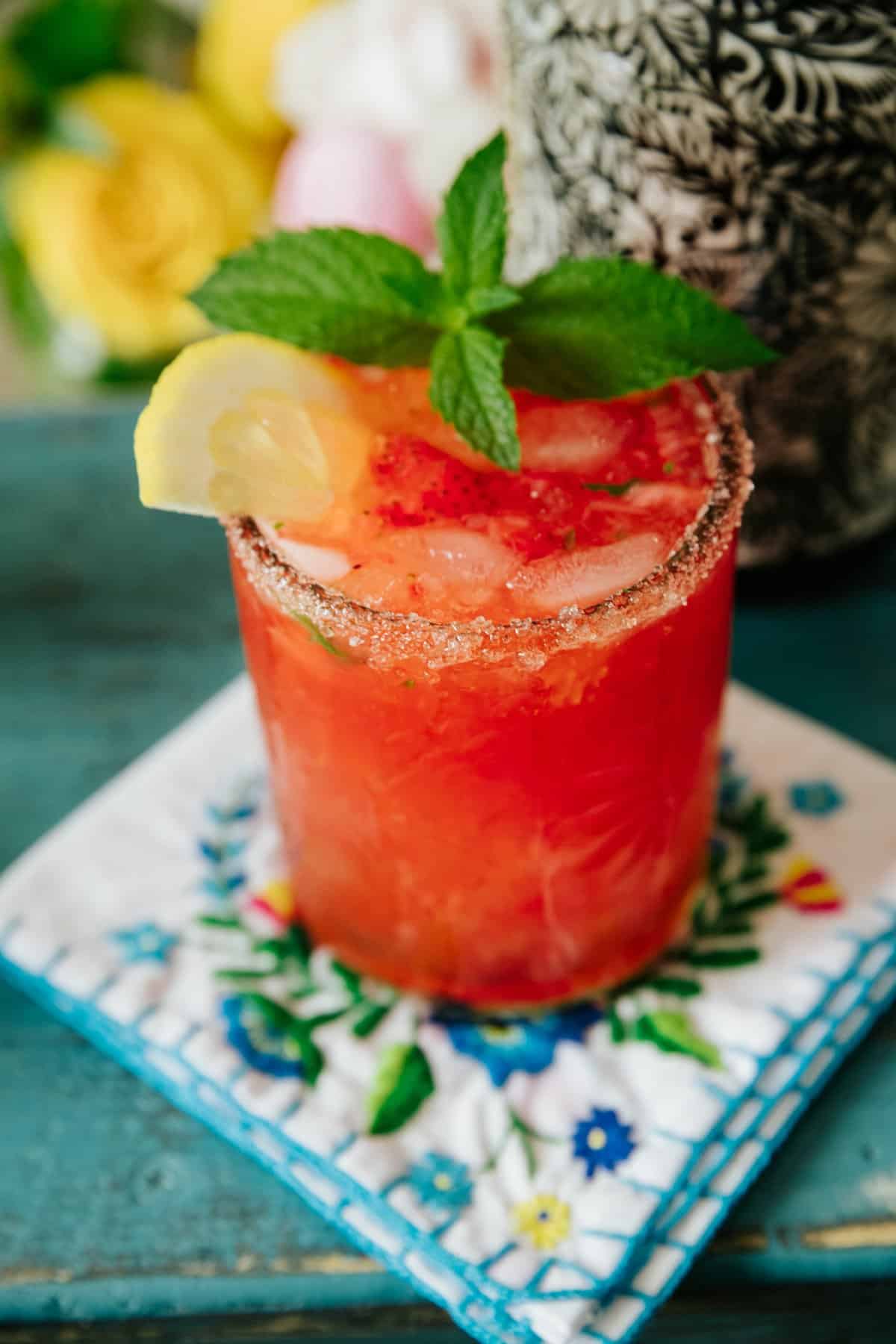 closeup of strawberry lemonade and tequila smash in a clear etched rocks glass with a sugar rim and garnished with a spring of fresh mint and a lemon wheel. 