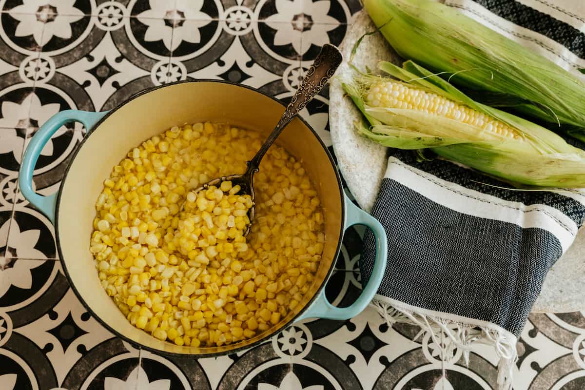 turquoise cast iron dutch oven with corn kernels and an antique silver spoon in it with 2 ears of corn off to the side. 