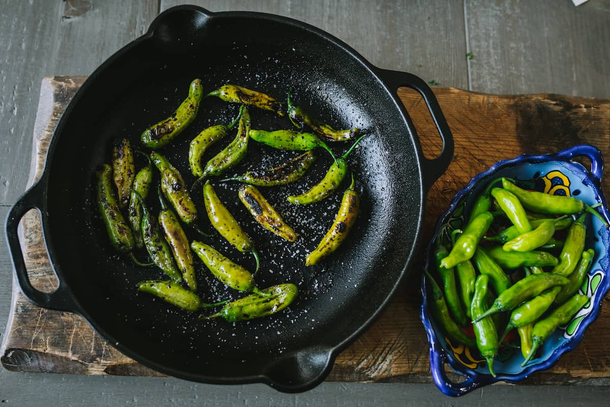 raw shishito peppers in a blue oval bowl and blistered shishito peppers in a cast iron skillet.