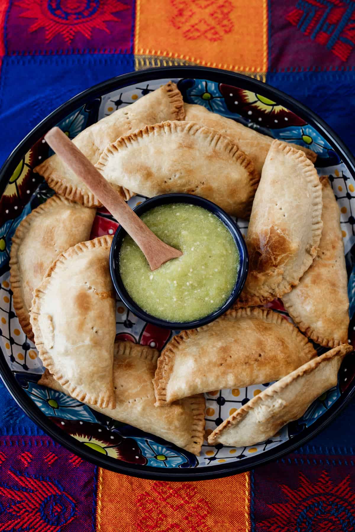 empanadas displayed on a Mexican talavera serving tray with a dish of salsa verde on top of a colorful Mexican tablecloth.