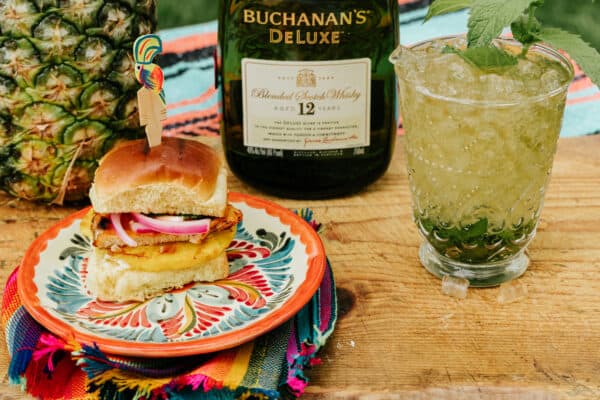 slider al pastor on a colorful plate with a bottle of buchanan's whisky and a whisky mint julep to the side.