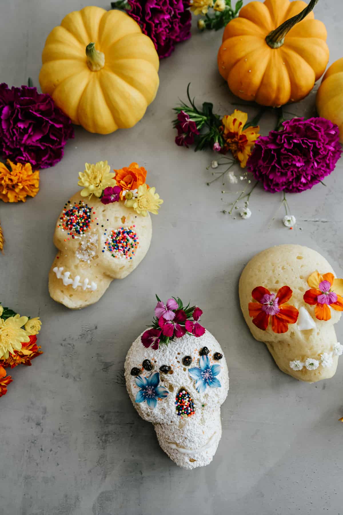 3 sugar skull cakes all decorated differently on a grey table with an assortment of pumpkins.