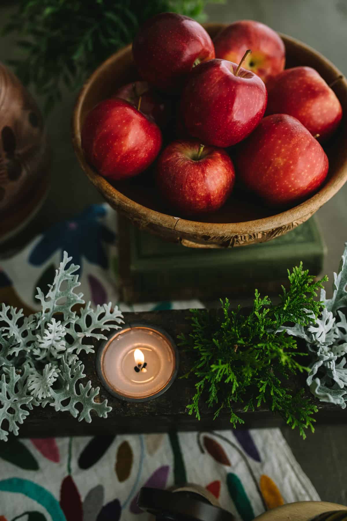 red apples in a carved wooden bowl next to wintry greenery and a candle.