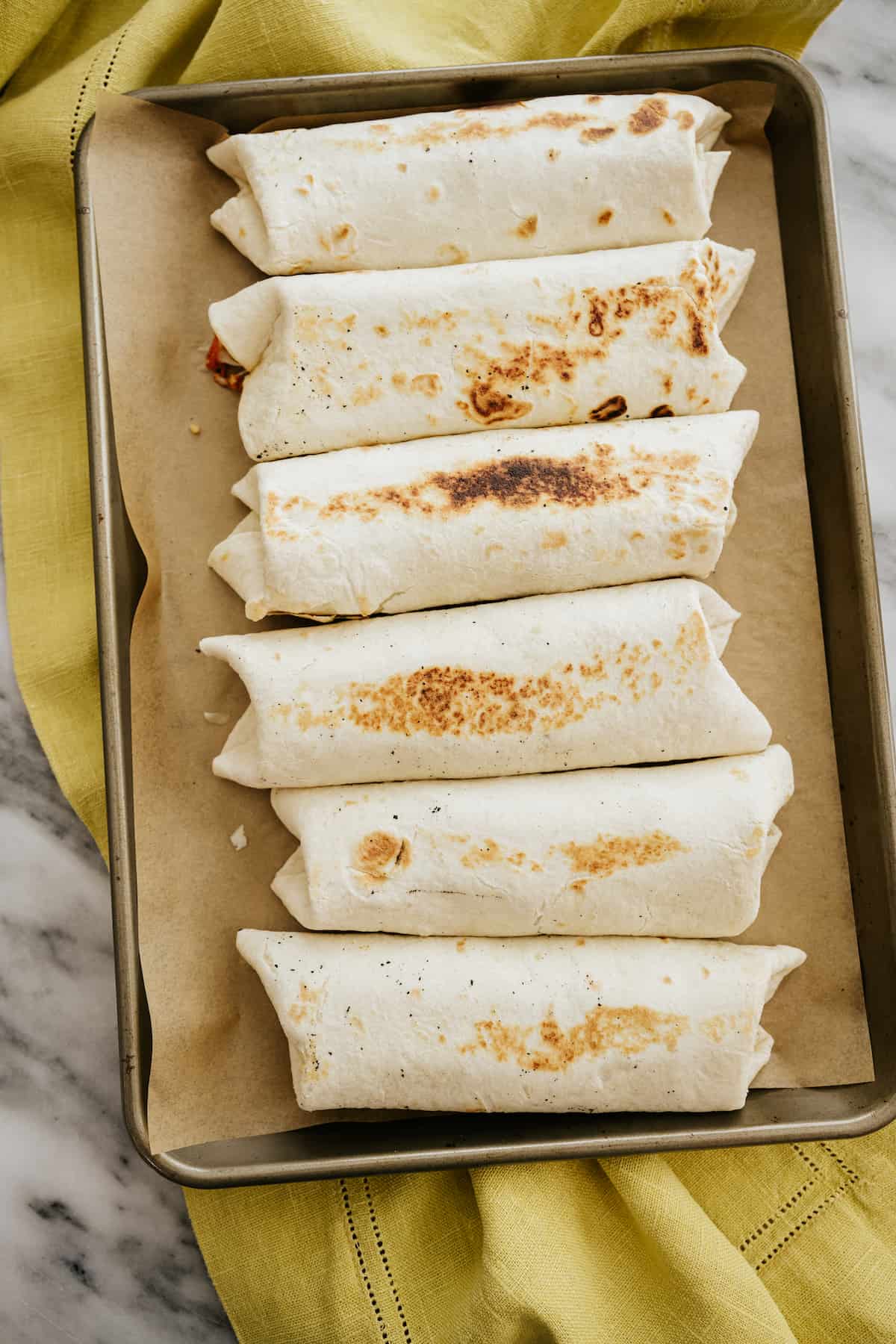6 wrapped and rolled breakfast burritos on a parchment-lined baking sheet.