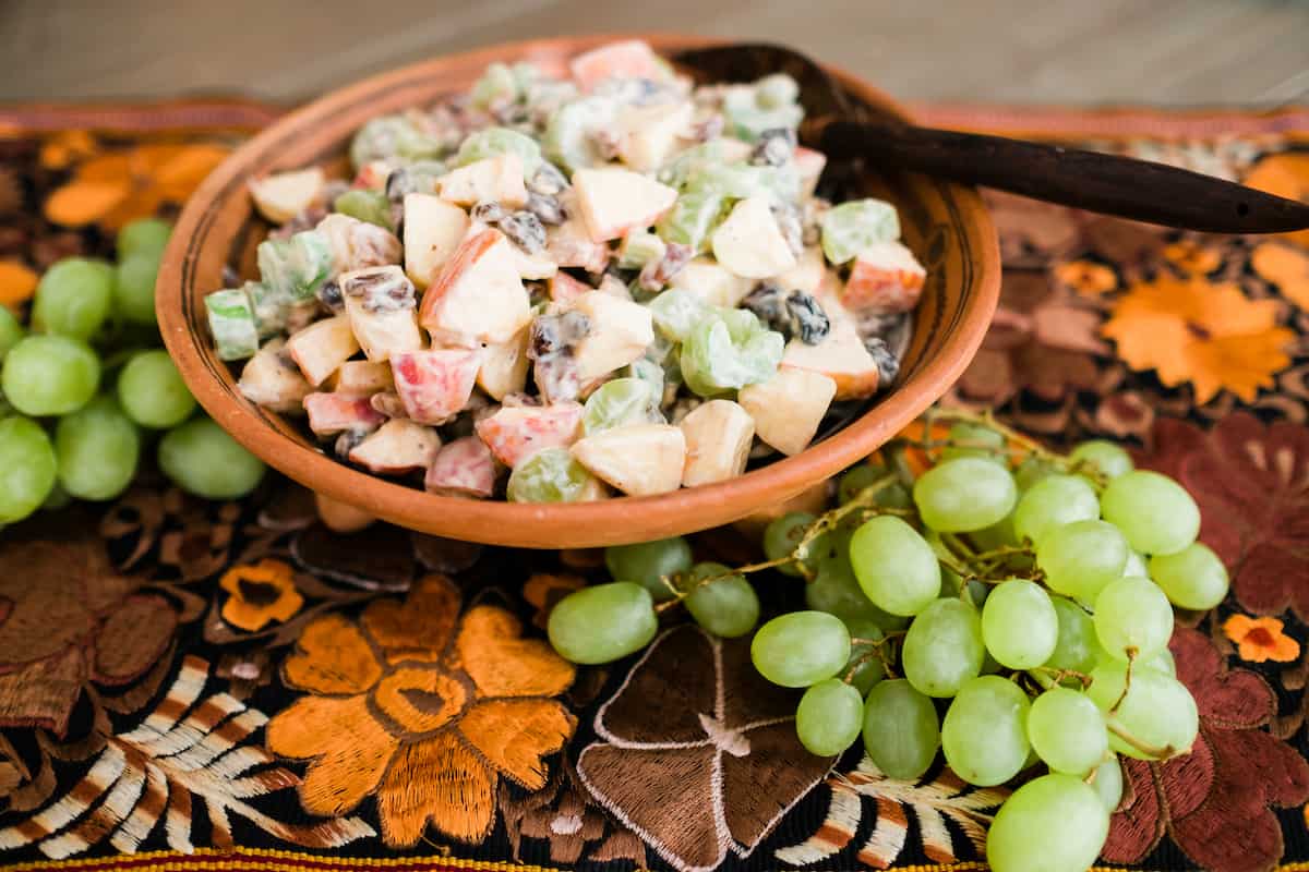 creamy waldorf salad with yogurt in a painted black and terracotta bowl with two bunches of green grapes on a thanksgiving inspired embroidered tablecloth.