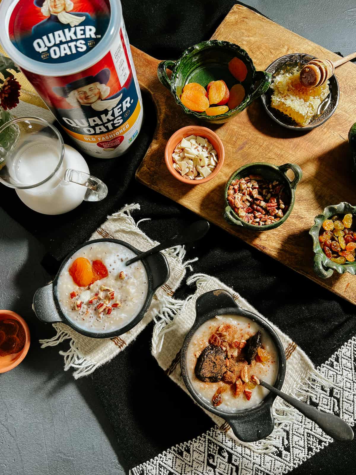overhead shot of a breakfast bar set up with bowls of dried fruits, chopped nuts, a pitcher of milk, a tin of quaker oats, and two cast-iron bowls of slow cooker oatmeal porridge.