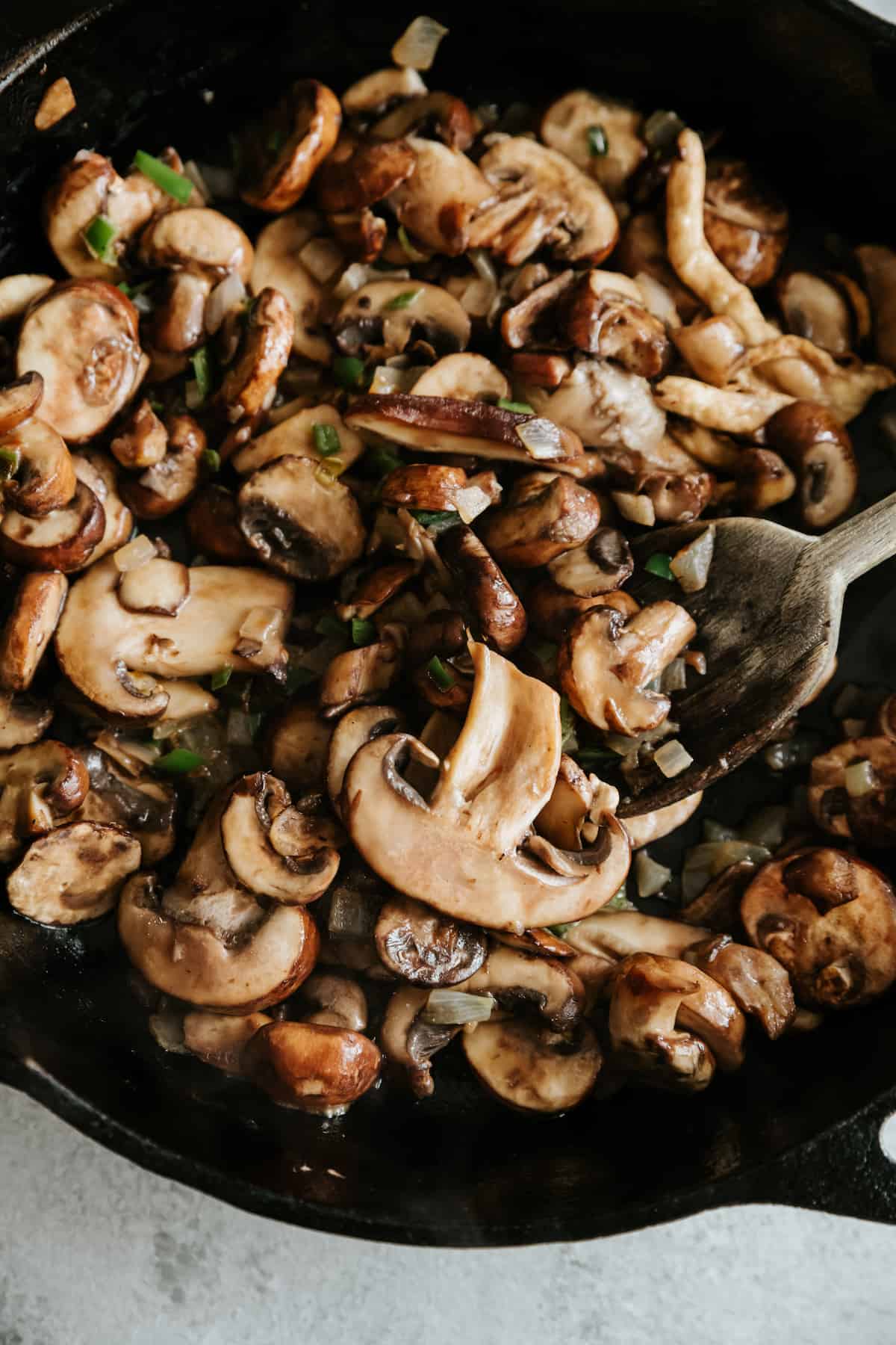 wooden spoon stirring a cast-iron pot filled with sautéed mixed mushrooms, peppers, and onions.