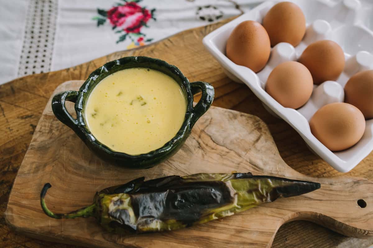 chile verde hollandaise in a metal bowl with handles on a wooden cutting board with a roasted green chile and a glass egg container.