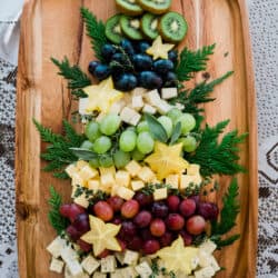 Tabla de quesos con forma de árbol de Navidad en una bandeja de madera, kiwi, uvas, carambola y queso rodeados de ramas perennes.