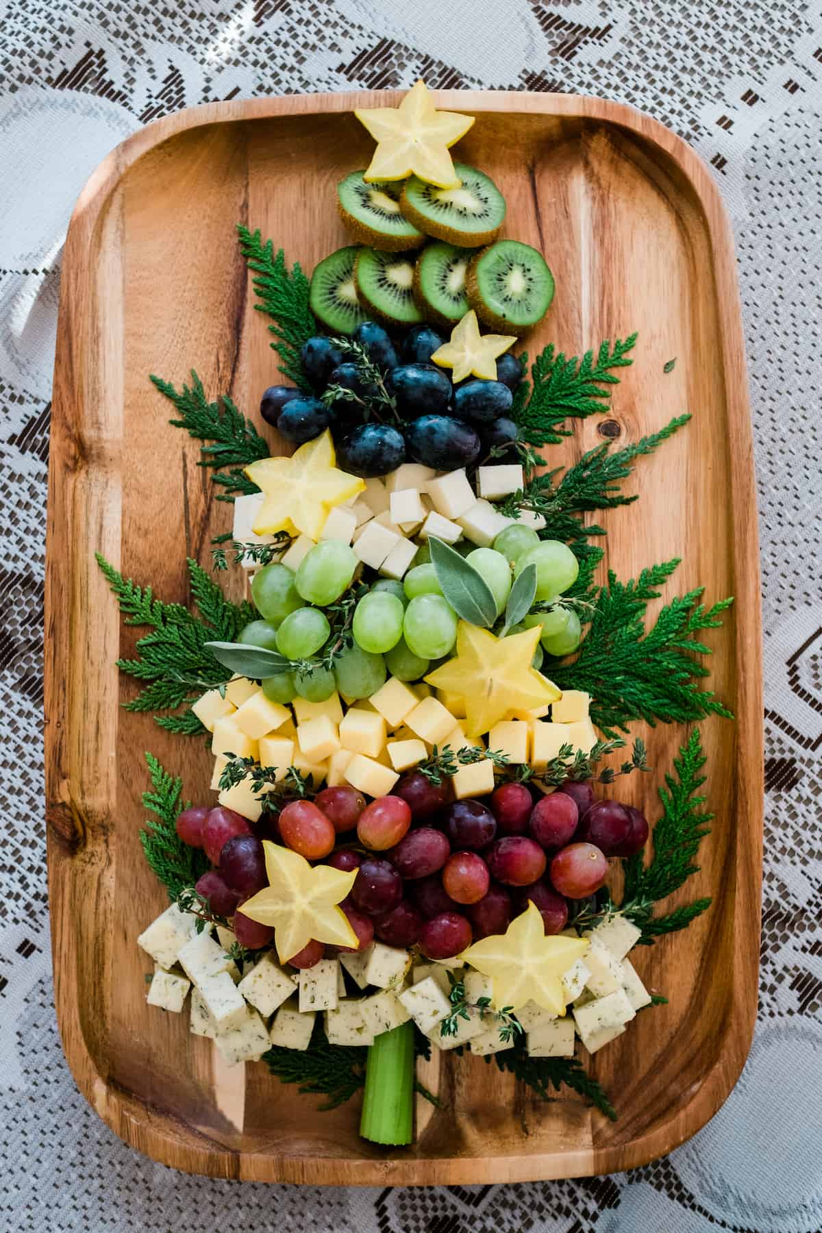 Tabla de quesos con forma de árbol de Navidad en una bandeja de madera, kiwi, uvas, carambola y queso rodeados de ramas perennes. 