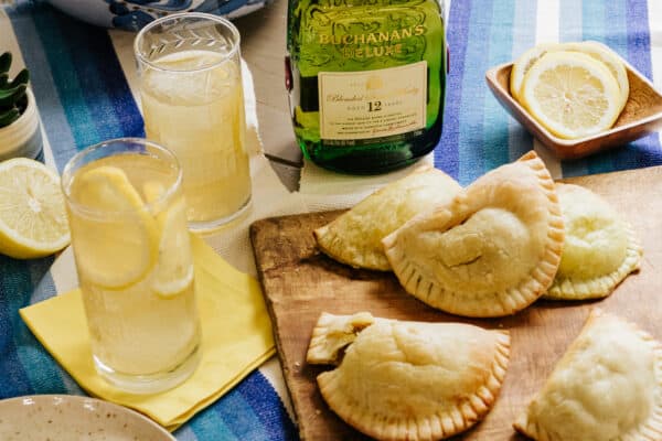 picadillo empanadas on a wooden cutting board next to two collins glasses filled with buchanan's whisky shandy cocktails.