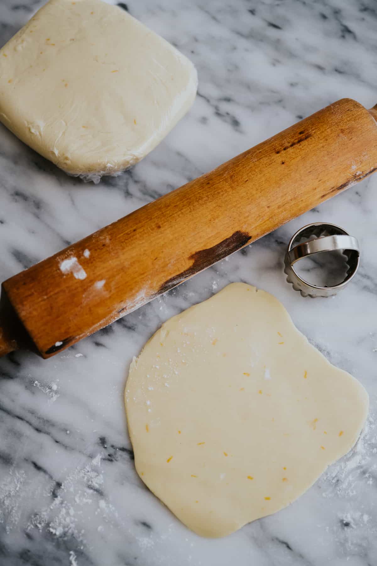 wooden rolling pin with rolled out shortbread dough and a scalloped cookie stamp on a marble surface.