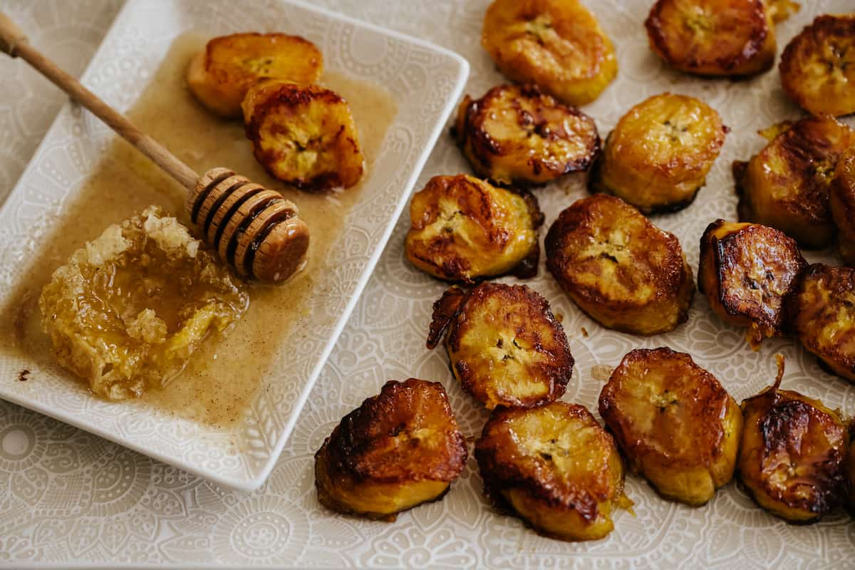 fried sweet plantains on a white rectangular serving platter with a smaller white plate with honeycomb and a honey drizzler stick.