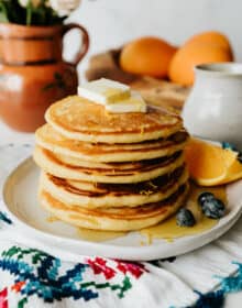 stack of 4 orange buttermilk pancakes on a white plate topped with maple syrup and fresh blueberries with a wedge of orange.