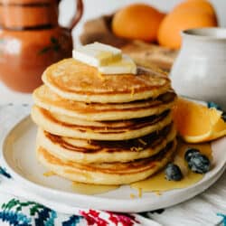 stack of 4 orange buttermilk pancakes on a white plate topped with maple syrup and fresh blueberries with a wedge of orange.