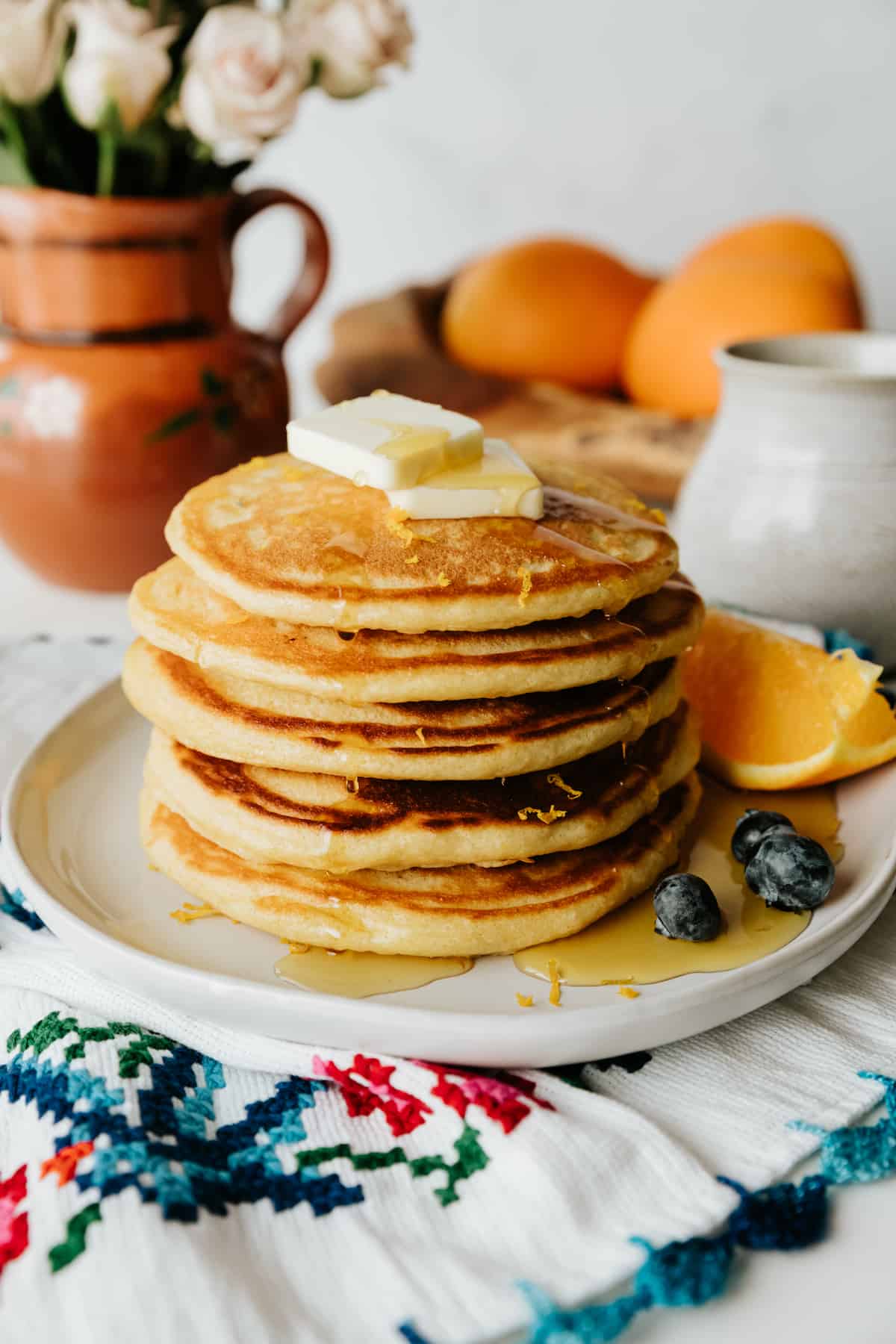 stack of 4 orange buttermilk pancakes on a white plate topped with maple syrup and fresh blueberries with a wedge of orange. 