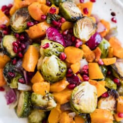 Serving platter of roasted butternut squash, burssels sprouts, red onions, and pomegranate seeds with a silver serving spoon.