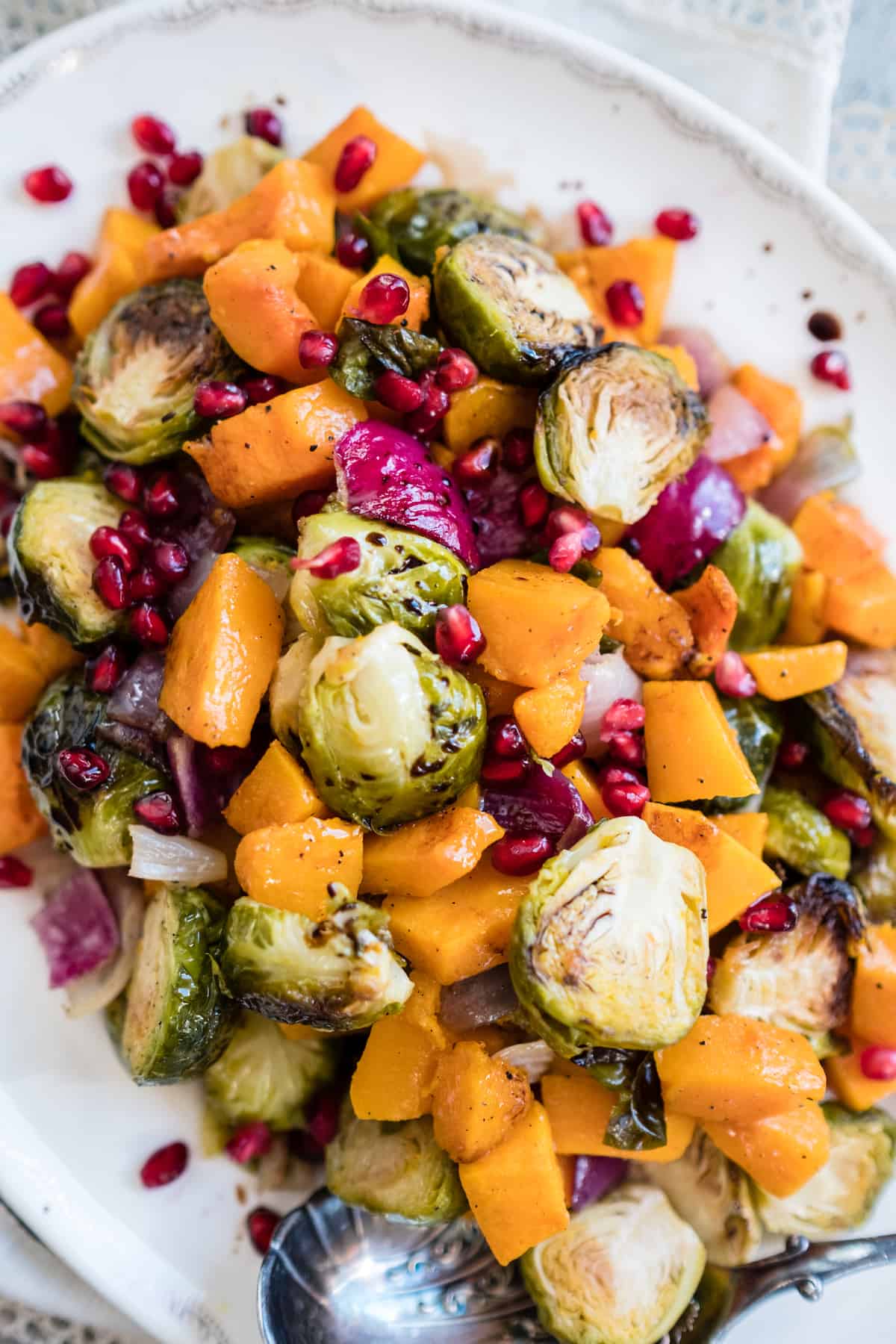 Serving platter of roasted butternut squash, burssels sprouts, red onions, and pomegranate seeds with a silver serving spoon.