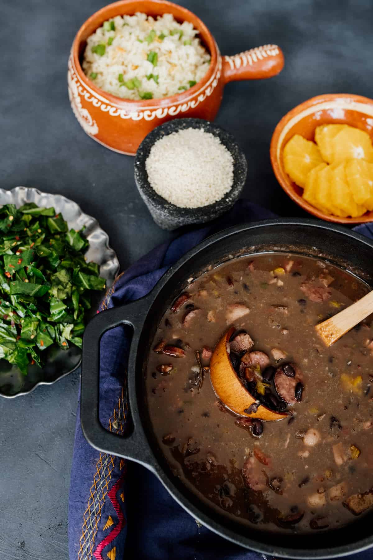 black cast iron dutch oven filled with Brazilian feijoada pork and black bean stew next to a serving bowl of steamed collard greens and a serving bowl of garlic butter rice.