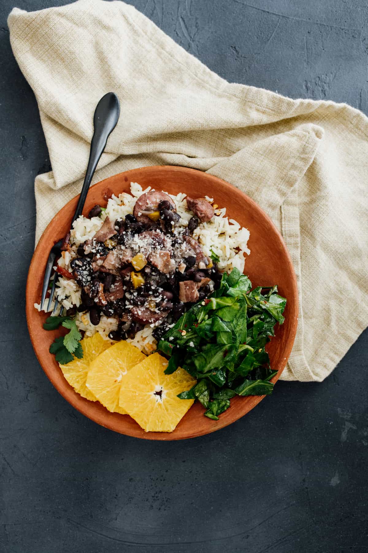 overhead shot of a terracotta bowl filled with steamed rice, Brazilian feijoada pork and black bean stew, orange segments and sautéed collard greens. 